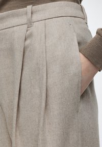 Gros plan sur un pantalon beige plissé avec la main dans la poche latérale, associé à une manche de pull en tricot marron visible au poignet.