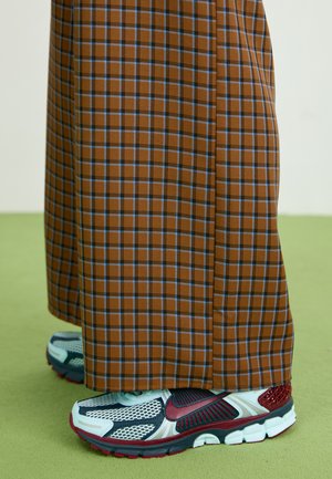 Pantalons larges à carreaux bruns avec des lignes bleues, associés à des baskets grises avec des accents rouges et un design en maille. Fond de sol texturé vert.