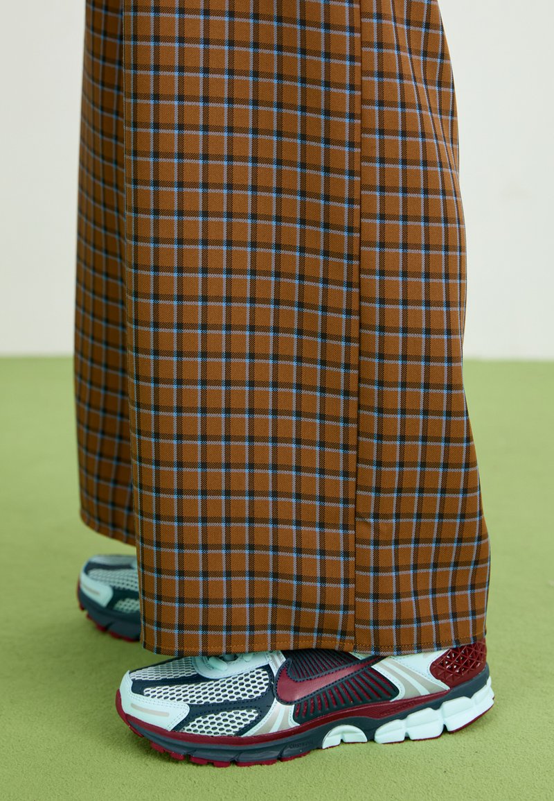 Bruine geruite wijde broek met blauwe lijnen, gecombineerd met grijze sneakers met rode accenten en een mesh-design. Groene gestructureerde vloer als achtergrond.