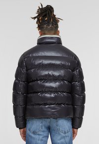 Zwarte pufferjas met glanzende, doorgestikte buitenkant; hoge kraag en elastische manchetten; gecombineerd met blauwe jeans, weergegeven vanaf de achterkant.