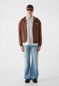 Rudas zipuotas džemperis su priekinėmis kišenėmis, vilkimas ant balto dryžuoto marškinėlio, derinamas su plačiais šviesiai mėlynais džinsais ir juodais sportbačiais.