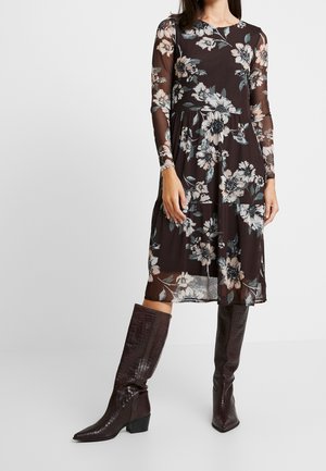 Vrouw draagt een donkervlinderige jurk tot op de knie met lange doorschijnende mouwen, gecombineerd met donkerbruine kniehoge textuurlaarzen.