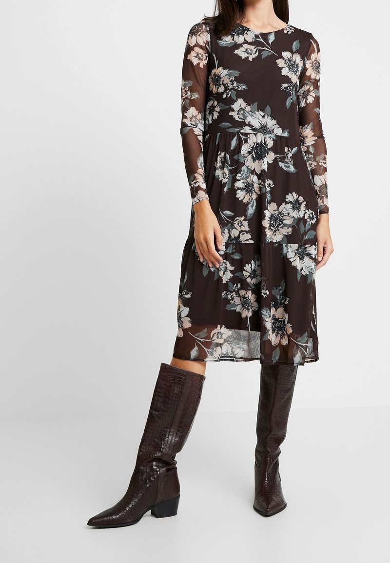 Femme portant une robe fleurie sombre mi-longue avec des manches longues transparentes, associée à des bottes marron foncé texturées montant jusqu'aux genoux.