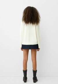 Biały dzianinowy sweter z prążkowanymi mankietami i krawędzią, zestawiony z ciemną mini spódnicą z denimu i wysokimi czarnymi kozakami. Widoczne kręcone włosy.