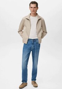 Beige lichtgewicht jack, witte t-shirt, lichte blauwe spijkerbroek en tan schoenen. Het jack heeft een rits aan de voorkant en een klassieke kraag.