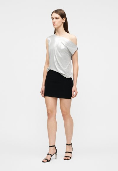 Γυναίκα που φοράει ασημί τοπ με έναν ώμο, μαύρη μίνι φούστα και μαύρα σανδάλια με τακούνια και λουριά, στέκεται μπροστά σε απλό φόντο.