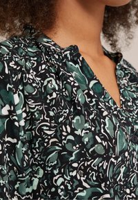 Zwarte stoffen blouse met een bloemenpatroon in groen en wit, met een V-hals, knopen aan de voorkant en geplooide schouderdetails.