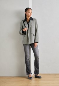 Blazer con estampado de pied de poule en gris y negro, combinado con una camisa negra de botones, jeans grises acampanados y zapatos planos negros, contra un fondo neutro.