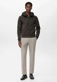 Sudadera con cremallera de color verde oscuro y capucha, combinada con pantalones claros de corte ajustado en gris y zapatos negros de cordones. Textura suave con diseño ceñido.