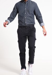 Chemise bleu foncé à boutons avec une poche poitrine, associée à un pantalon cargo noir avec des poches latérales, et des baskets blanches.
