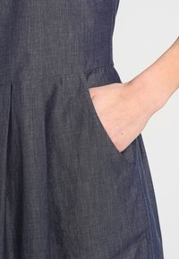 Dunkelblaues Denim-Kleid mit Taschen, das einen taillierten Oberteil und einen ausgestellten Rock hat. Der Stoff hat eine leichte Textur und eine weiche Oberfläche.
