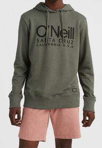 Felpa verde oliva con stampa nera davanti, dotata di cordini e polsini. Abbinata a pantaloni corti rosa chiaro.