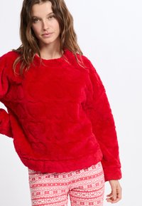 Červený plyšový svetr s motivem srdce, v kombinaci s bílými vzorovanými kalhotami se sněhovými vločkami a dekoračními pruhy.