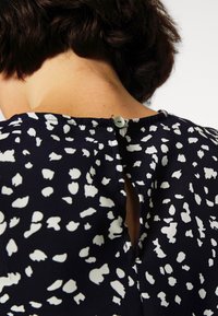 Laivastonsininen blouse, jossa on valkoisia abstrakteja pilkkuja, takana nappikiinnitys ja avaus avainreijällä. Sujuva pinta ja rento malli.