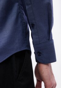 Κοντινό πλάνο του χεριού ενός ατόμου που φοράει μπλε πουκάμισο με υφή και μανσέτα με κουμπί, πάνω από μαύρο παντελόνι, σε λευκό φόντο.