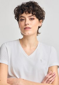 Witte V-hals t-shirt van zacht materiaal, met korte mouwen en een klein geborduurd logo op de borst. Gladde textuur.
