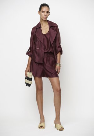 Femme portant une veste en satin bordeaux, un débardeur et un short, tenant une petite pochette rayée, avec des sandales à talons tissées beige et des bracelets en or.