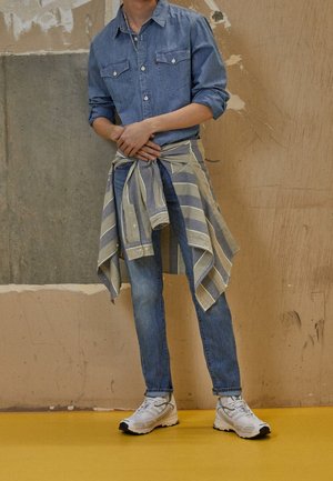 Person iført opkrøllede denimskjorte og jeans med en stribet skjorte bundet omkring livet, stående op ad en struktureret væg på et gult gulv.