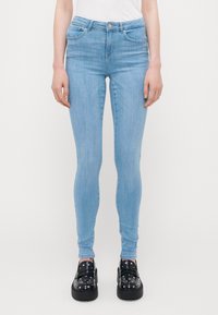 Persona que lleva puesta una camiseta azul claro y unos loafers de plataforma negros con tachuelas, de pie contra un fondo blanco liso.