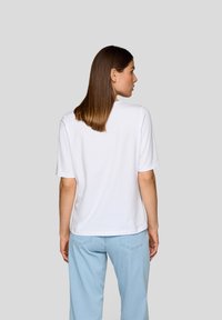 White cotton t-shirt with short sleeves, crew neck, and a relaxed fit, paired with light blue denim pants, viewed from the back.