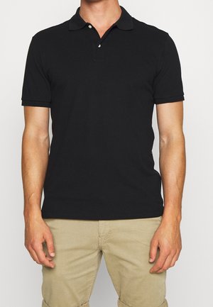 Mann trägt ein eng anliegendes schwarzes Kurzarm-Poloshirt und beige Hosen, steht mit entspannten Armen vor einem schlichten Hintergrund.