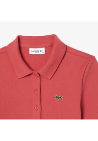 Polo de corte slim en coral suave con canalé, con un placket de tres botones y el logo del cocodrilo verde de Lacoste en el lado izquierdo del pecho.