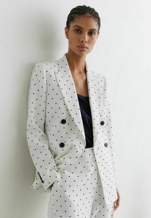 Frau trägt weißen doppelt geknöpften Blazer und passende Hose mit schwarzen Polka-Dots, steht mit den Händen in den Taschen vor einer weißen Wand.