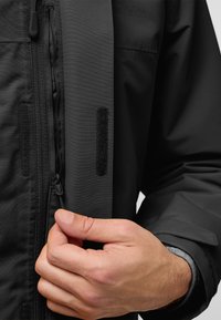 Zwarte waterdichte jas met een gestructureerde stof, een ritssluiting aan de voorkant en klittenbandaccenten. De hand grijpt de ritsstrop.