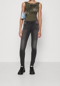 Grå skinny jeans med en blekt textur, kombinerat med en mörkgrön ribbad linne med tryckt text. Accessoarer med en liten blå handväska.