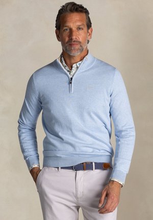 Lichtblauwe pullovertrui met een ritssluiting bij de hals, geribbelde manchetten en zoom, gelaagd over een bloempatroon shirt, gecombineerd met lichte broek.