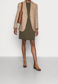 Blazer in lino beige su un abito verde oliva, abbinato a stivaletti in pelle marroni e una borsa a tracolla coordinata. Texture morbide e vestibilità su misura.