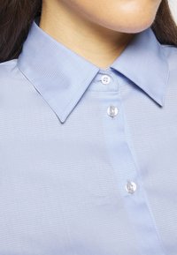 Camicia azzurro chiaro con bottone, caratterizzata da un tessuto testurizzato, colletto classico e bottoni trasparenti, con un sottile motivo a griglia.
