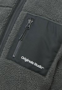 Jakna od flisa u tamno sivoj boji s teksturiranom tkaninom. Ima džep sa patentnim zatvaračem i "Originals Studio" logo u bijeloj boji na glatkoj površini.