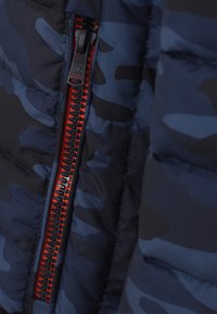 Närbild av en marinblå camouflagemönstrad pufferjacka med en delvis zippad röd och svart plastdragkedja.