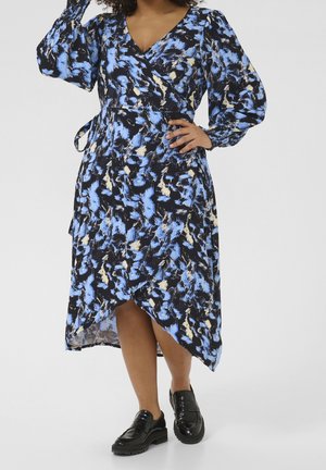 Woman wearing a blue, black, and beige patterned wrap dress with long puffed sleeves and black chunky shoes, posing with hand on hip.