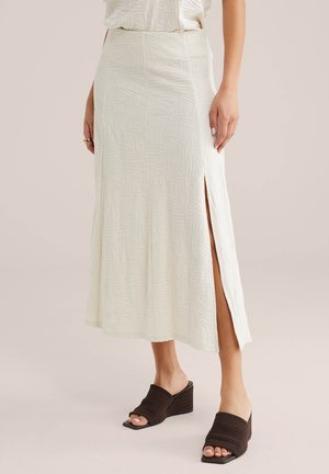 Frau trägt einen cremefarbenen, strukturierten Midirock mit Seitenschlitz und schwarzen, geflochtenen Keilsandalen mit offenem Zehenbereich, steht vor einem einfarbigen Hintergrund.