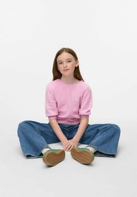 Ragazzina con lunghe capelli castani seduta a gambe incrociate, vestita con un maglione rosa, jeans blu, calze bianche e scarpe verdi su uno sfondo bianco.