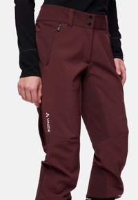Nahaufnahme einer Person, die bordeauxfarbene Outdoor-Hosen mit Reißverschlusstaschen und dem Vaude-Logo auf dem Oberschenkel trägt, kombiniert mit einem schwarzen Langarmoberteil.