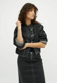 Frau mit lockigem, dunklem Haar, die eine schwarze Jeansjacke mit silberfarbigen Verschlüssen und einen passenden, hoch taillierten Rock trägt, schaut seitlich.