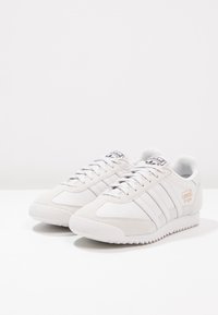 Weiße Sportsneaker mit grauen Wildlederakzenten, ausgestattet mit einem Text-Obermaterial, drei weißen Streifen und einer strukturierten Gummisohle. Logo an der Seite.