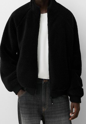 Black fleece jacket with a zip-front, high collar, and ribbed cuffs. The fabric has a soft, textured finish. Worn over a white shirt.