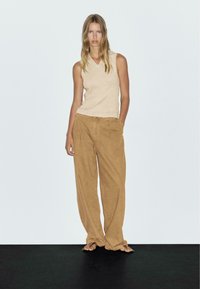 Woman with long blonde hair wearing a sleeveless beige top, loose brown pants, and black flip-flops standing against a plain white background.