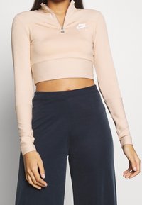 Ljus persikofärgad kortärmad topp med halv dragkedja och ribbad textur, samt en vit Nike-logotyp på den övre vänstra sidan. Tillsammans med mörka byxor.