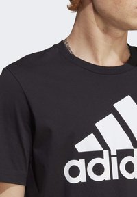 Černé bavlněné tričko s kulatým výstřihem a krátkými rukávy, se výrazným bílým logem Adidas a designem tří diagonálních pruhů.