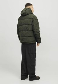 Dunkelgrüne Steppjacke mit Kapuze, die gestickte Abschnitte und eine strukturierte Oberfläche aufweist. Kombiniert mit lockeren schwarzen Cargo-Hosen.