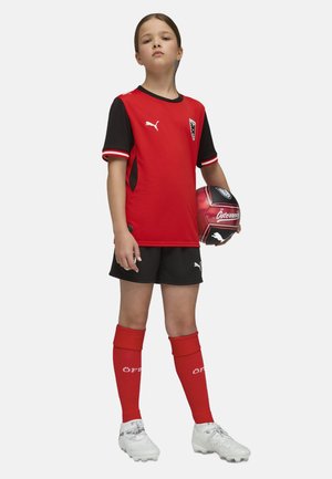 Niño con uniforme de fútbol rojo y negro sosteniendo un balón de fútbol rojo y negro, de pie con confianza frente a un fondo blanco.