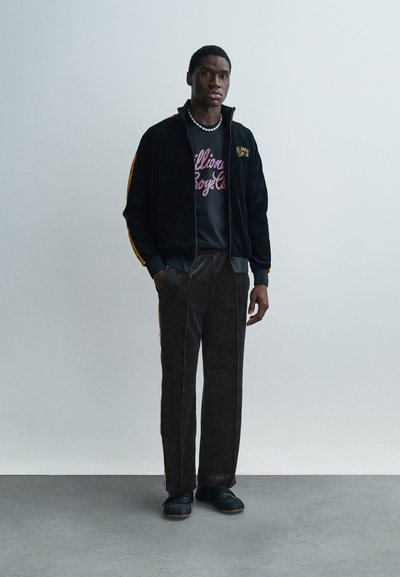 Μαύρο βελούδινο ζακέτα με φερμουάρ και κίτρινες ρίγες, γκρι μπλουζάκι με γραφικό σχέδιο, σκούρο παντελόνι από κοτλέ, και μαύρα αθλητικά παπούτσια με καφέ σόλες.