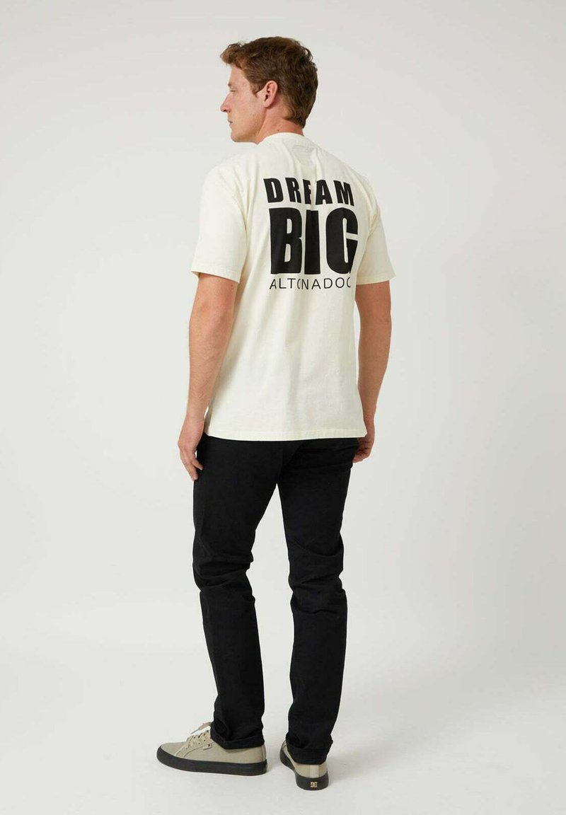 Camiseta color crema con la frase "SUEÑA EN GRANDE" en letras negras y gruesas en la parte trasera. Se lleva con pantalones negros y zapatillas beige, mirando ligeramente hacia la izquierda.