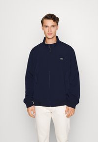 Mornarska zip-up jakna z visokim ovratnikom, lahkotna tkanina, stranska žepa in majhnim zelenim logotipom na prsih; elastični manšeti in rob.
