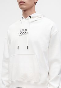 Jeune personne portant un sweat à capuche blanc avec des cordons noirs et un logo texte noir sur la poitrine.